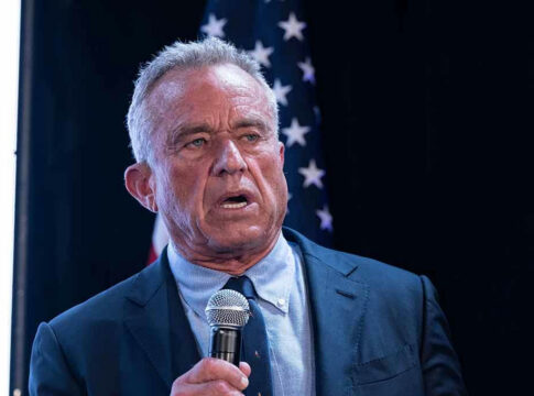 Man in suit holding a microphone, American flag background.
